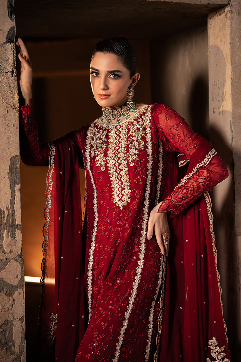 Woman in a red traditional outfit with intricate white embroidery standing against a textured wall.