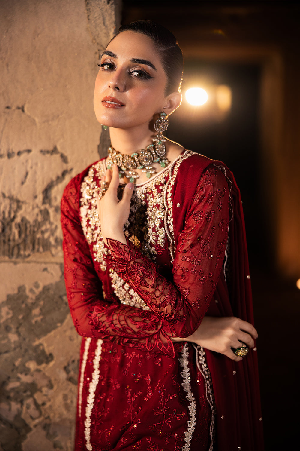 Woman in a red traditional outfit with intricate designs and jewelry against a textured wall.