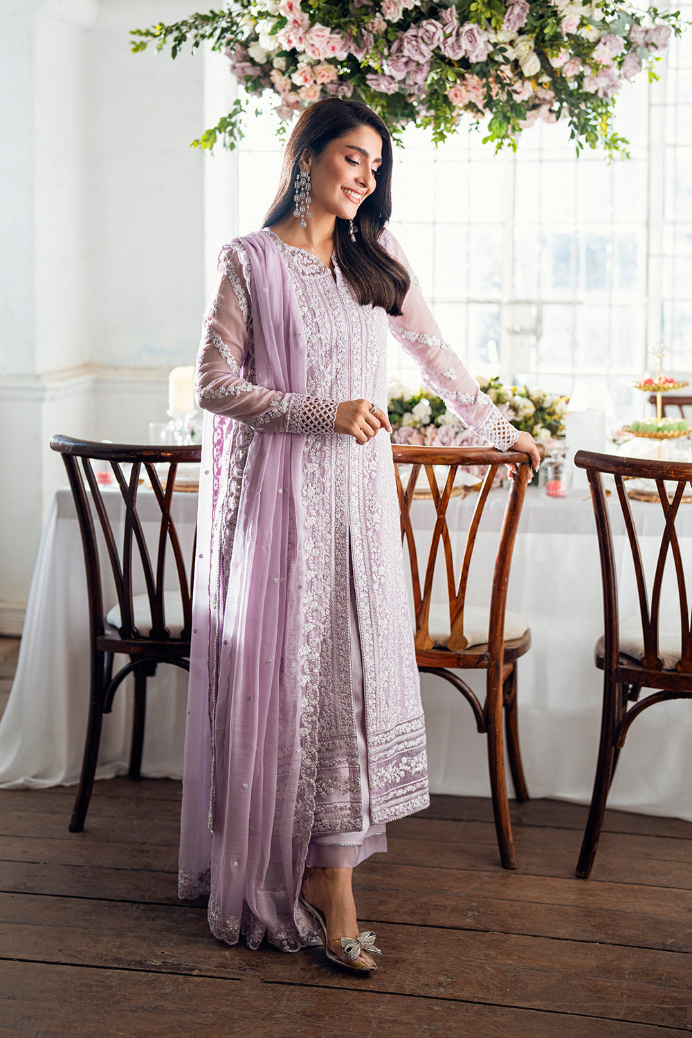 Woman in a light purple and white traditional outfit standing in a decorated room with flowers and a table.