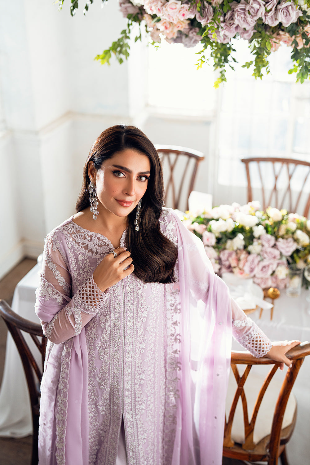Woman in a light purple and white traditional outfit standing in a decorated room with flowers and furniture.