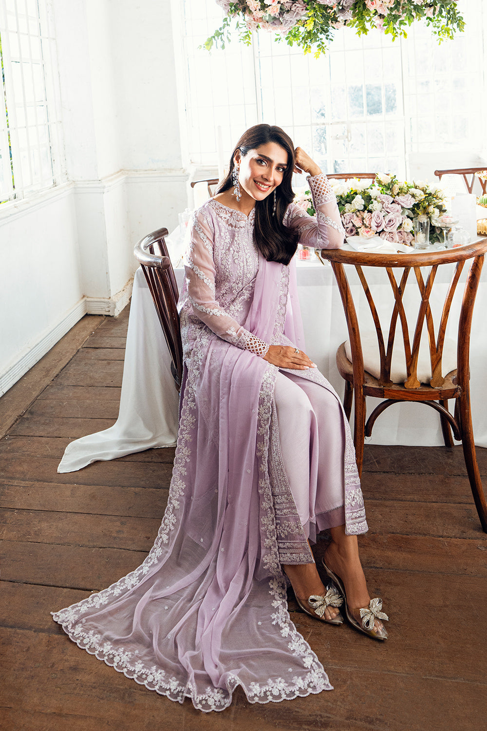 Woman in a pink lace dress sitting at a wooden table with flowers in the background