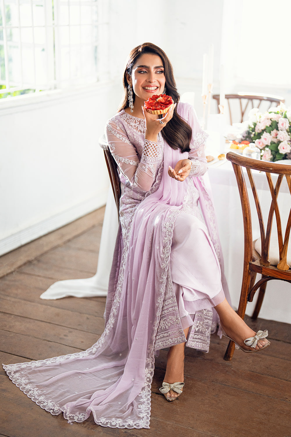 Woman in a pink saree holding a flower, sitting on a wooden chair indoors.
